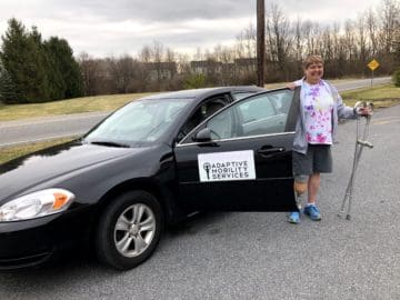 Person with right leg amputation standing next to black Adaptive Mobility sedan, smiling and holding hand crutches.