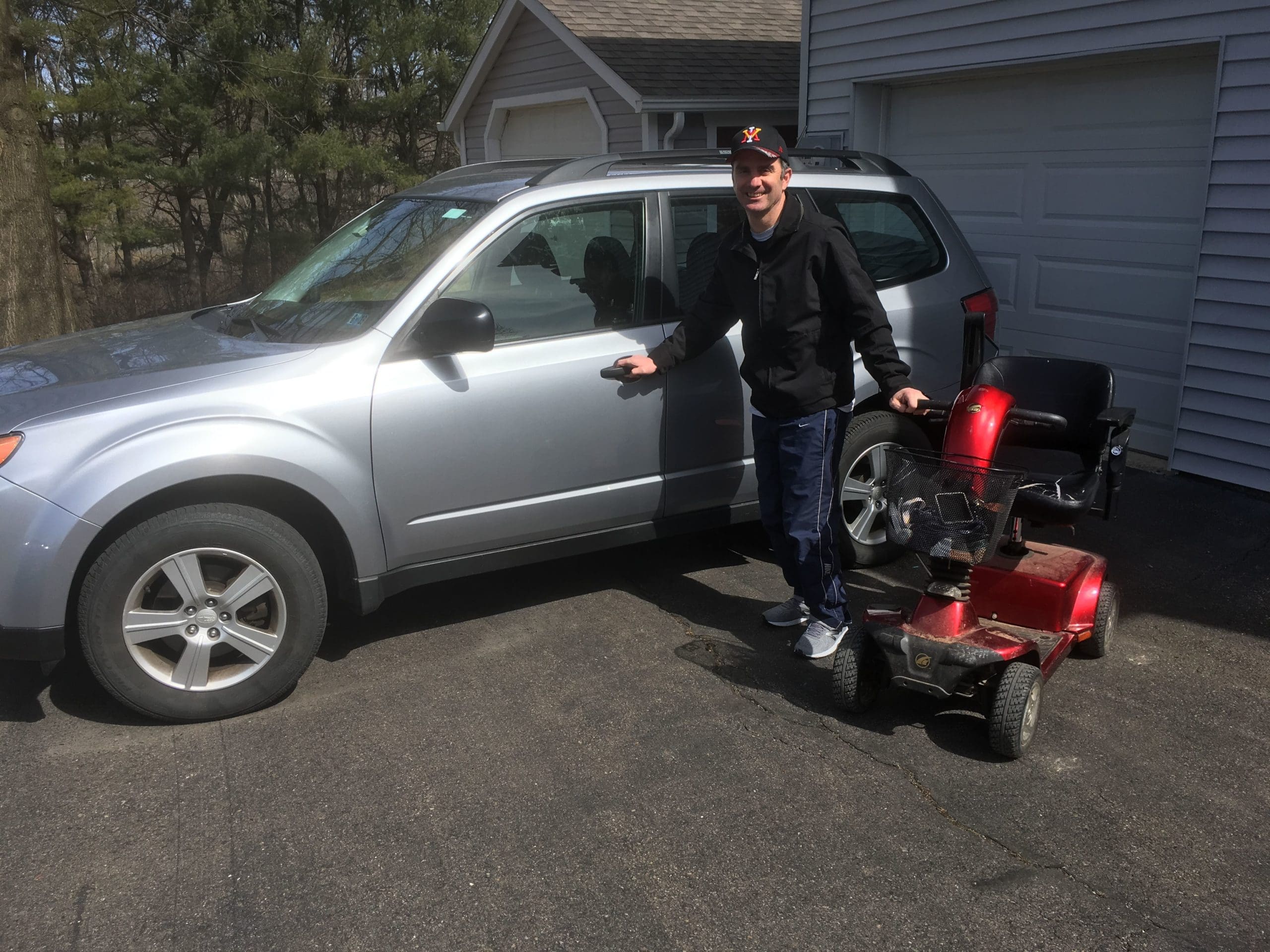 Person standing next to silver SUV, holding the door handle with one hand and the other resting on the handle of his motorized wheelchair scooter.