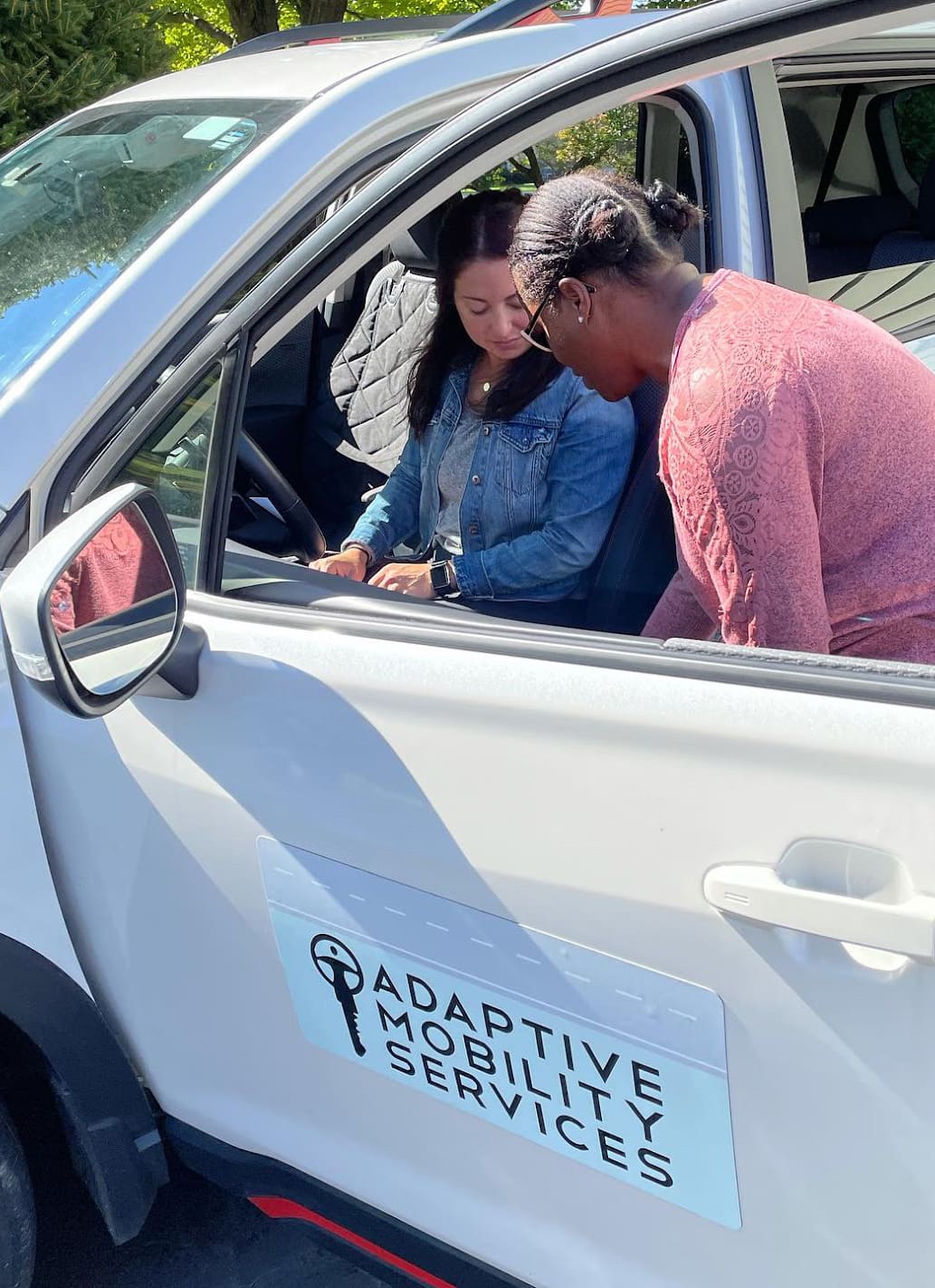 Driver Assessment instructor showing an OT, who is sitting in the driver's seat of a white SUV, the different adaptive equipment inside the vehicle.