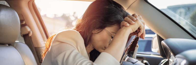 A distressed woman is sitting in a car. She has her head and hands resting on the steering wheel.